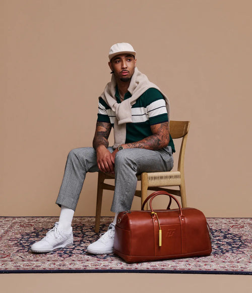 Man sitting on a chair with a brown leather duffel bag next to him against a beige wall.