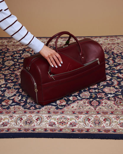 Red leather duffel bag on a patterned rug with a hand reaching towards it.